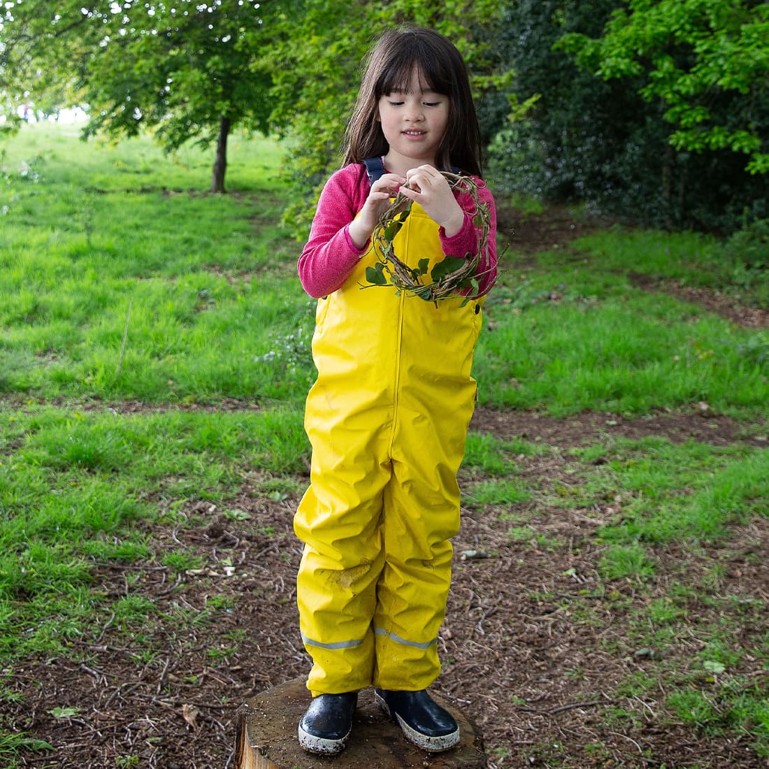A girl in Puddleflex Waterproof Fleece Lined Dungarees Yellow stands on a tree stump holding a leafy crown in a woodland setting.