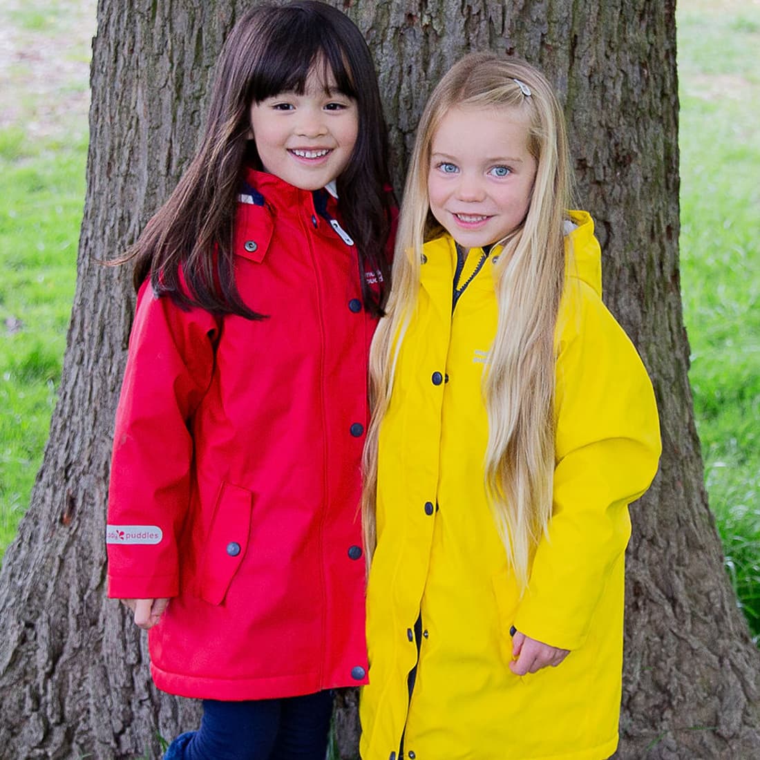 Two girls in raincoats stand by a tree. One wears a red Puddleflex Waterproof Fleece Lined Jacket. The other wears yellow.