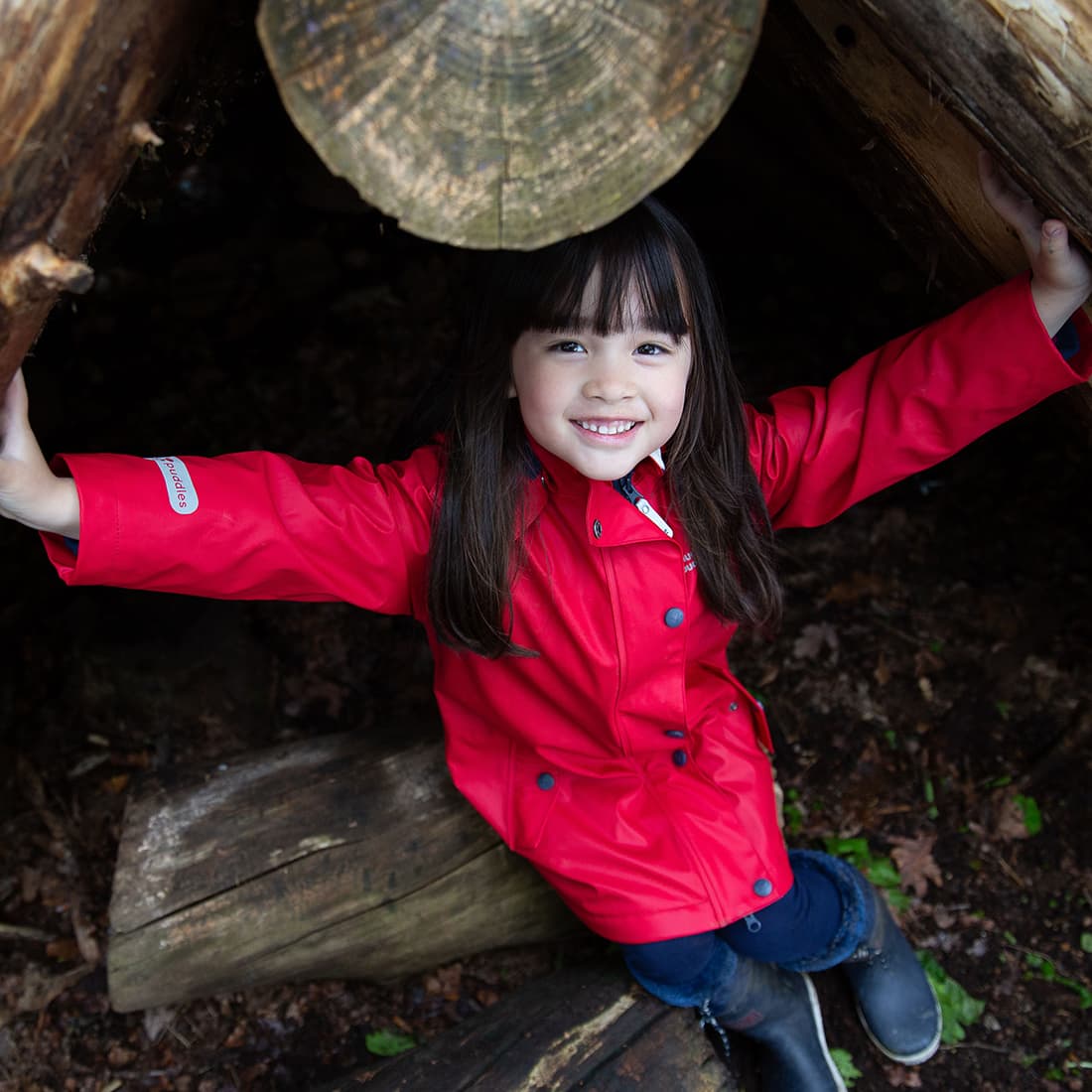 A girl smiles in a den wearing a red Puddleflex Waterproof Fleece Lined Jacket, jeans and black boots.