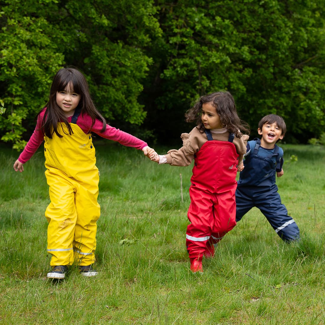 Three children play in a field wearing Puddleflex Waterproof Fleece Lined Dungarees in yellow, red, and navy. They're holding hands and laughing.