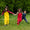 Three children play in a field wearing Puddleflex Waterproof Fleece Lined Dungarees in yellow, red, and navy. They're holding hands and laughing.