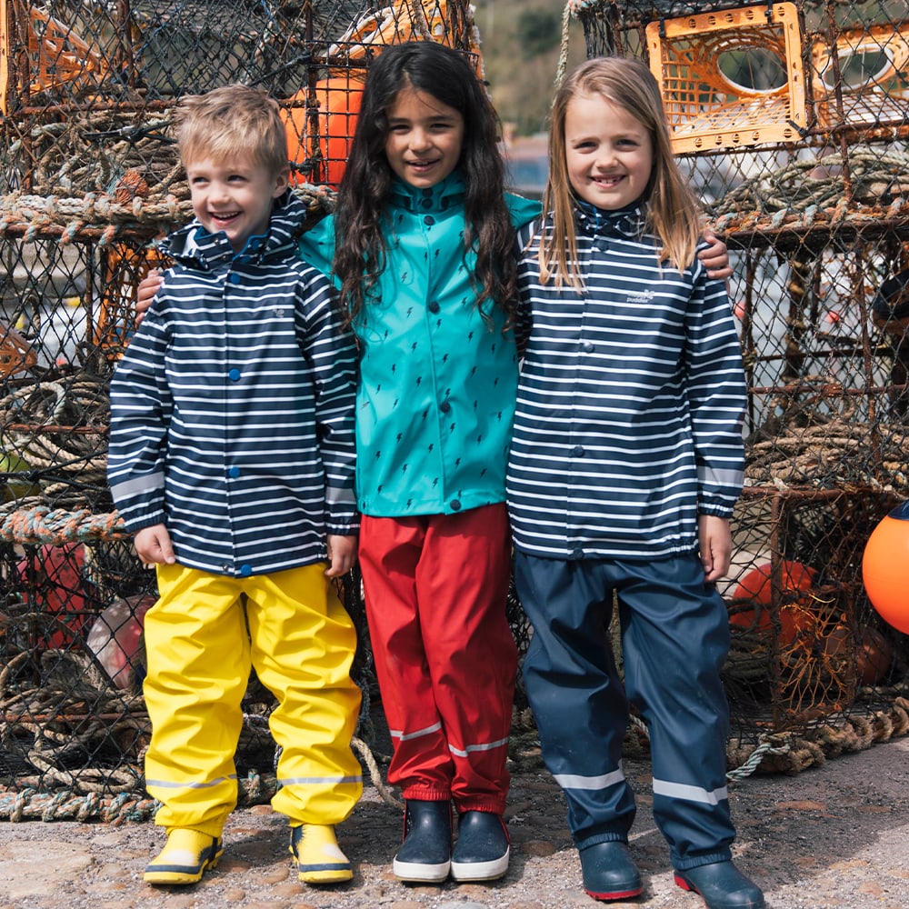 Three children wear Puddleflex Waterproof Fleece Lined Trousers in yellow, red and navy. They are stood in front of fishing nets.