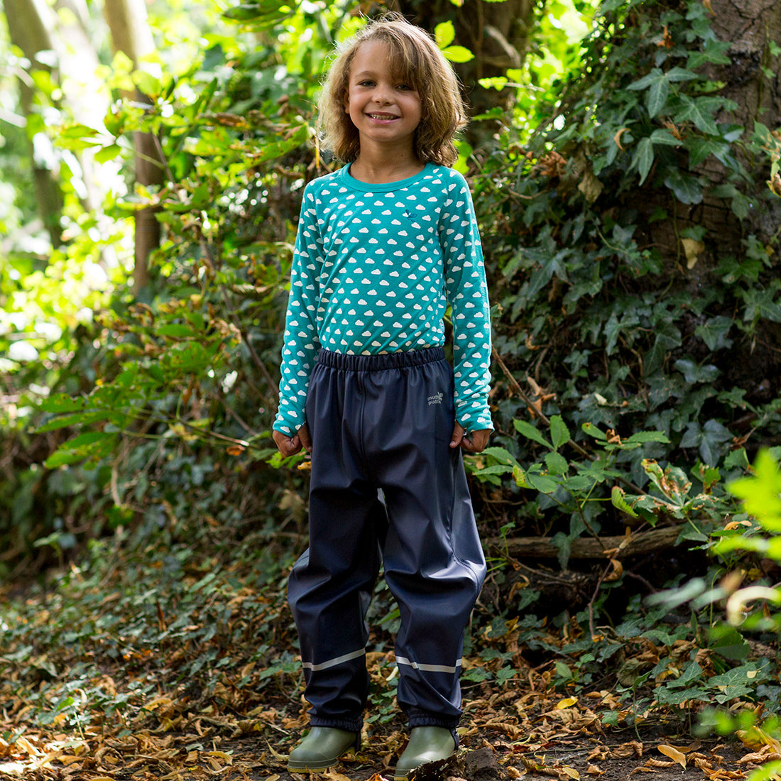 A young child stands in the woods wearing Rainy Day Waterproof Trousers Navy, and a patterned top and green wellies.