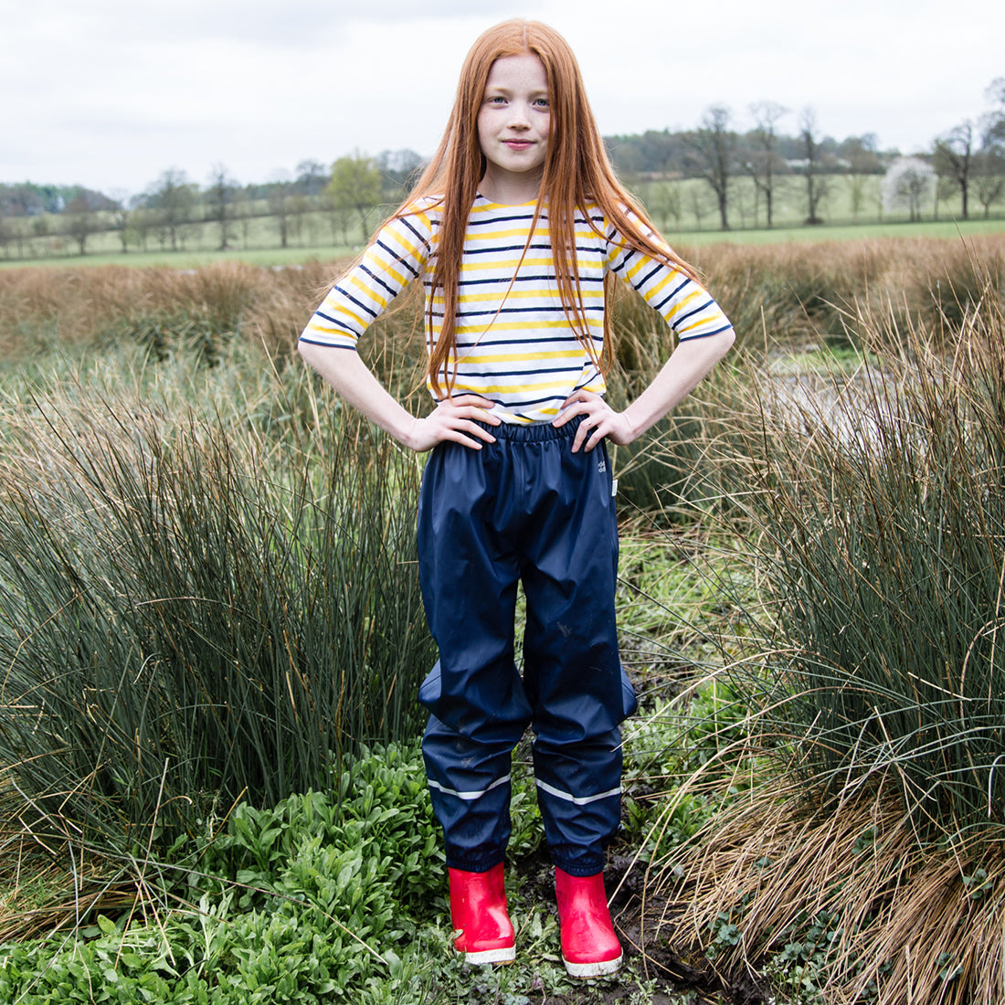 A girl stands in a grassy field wearing Puddleflex Waterproof Fleece Lined Trousers in navy with a striped top and red wellies.