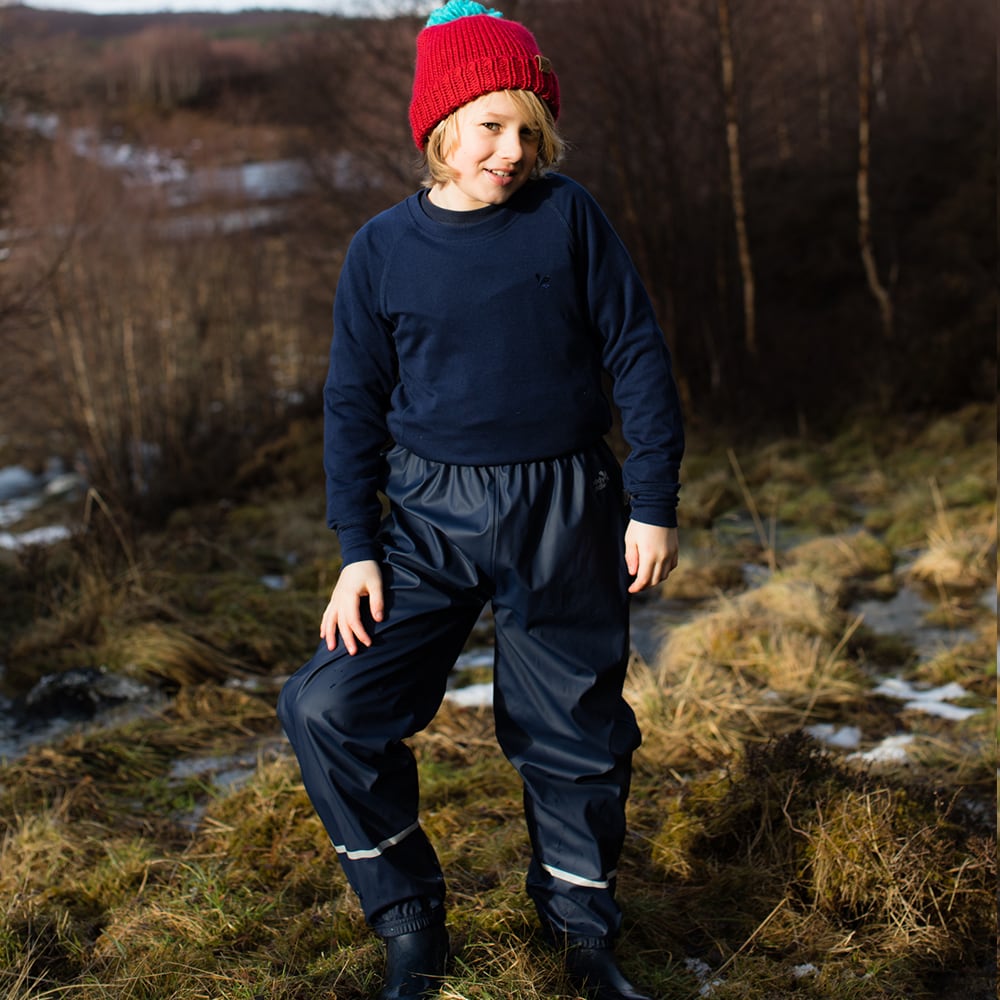 A boy standing outdoors wearing Rainy Day Waterproof Trousers Navy, a navy jumper and a red bobble hat.