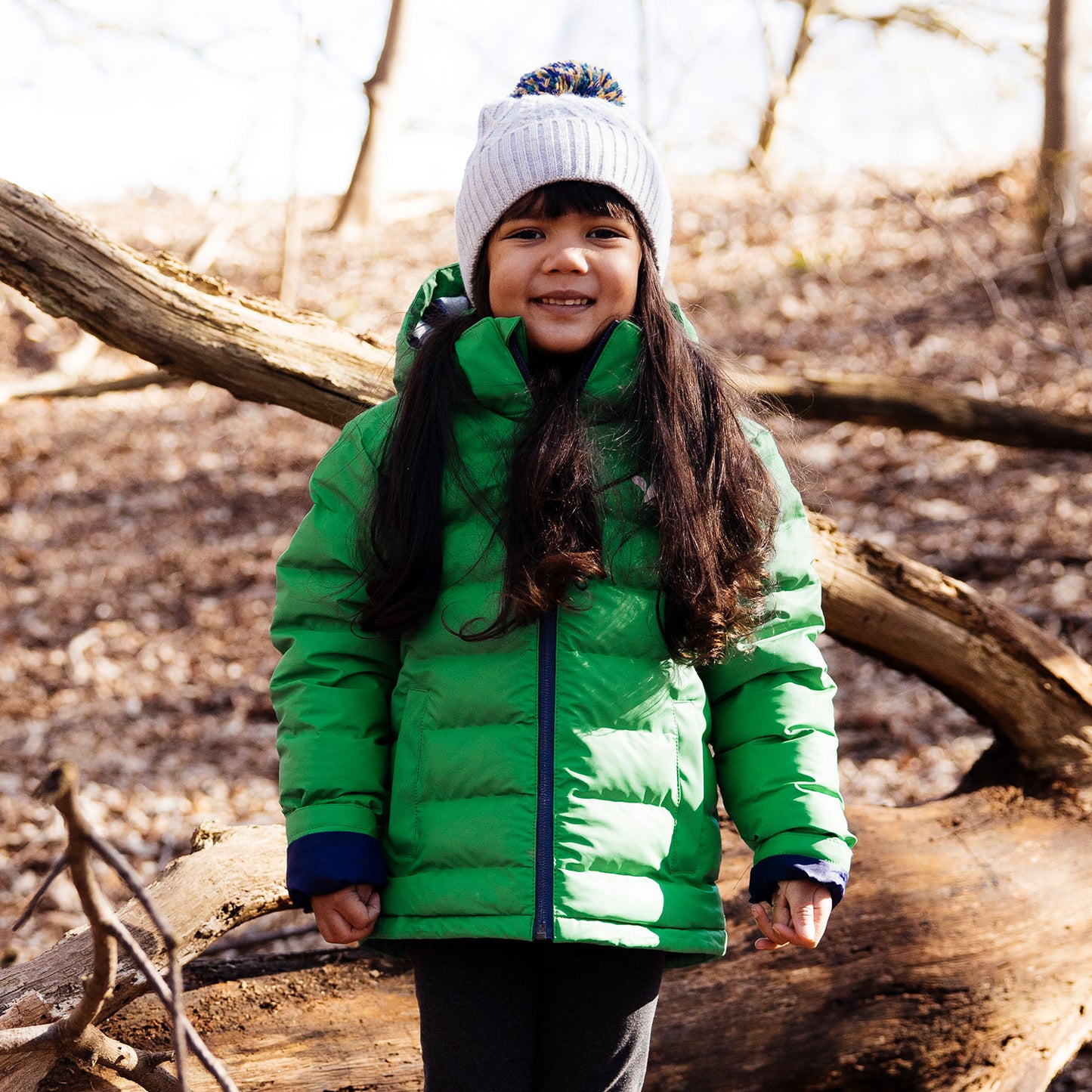 A smiling child wearing a green waterproof puffer jacket and a white hat, standing on a fallen tree trunk in a forest setting.