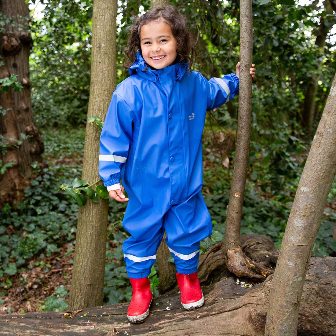 A young child wears a 'Rainy Day Waterproof Puddle Suit Blue' while standing amongst trees, with red wellington boots on.