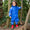 A young child wears a 'Rainy Day Waterproof Puddle Suit Blue' while standing amongst trees, with red wellington boots on.