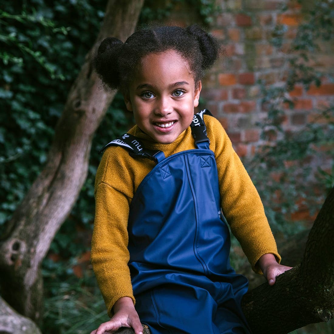A smiling child in Rainy Day Waterproof Dungarees Navy sits on a tree branch against a leafy, brick wall backdrop.