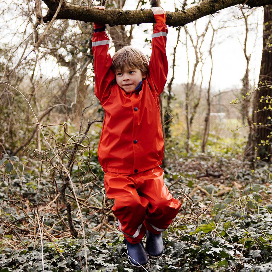 A child wearing a Rainy Day Waterproof Jacket and Dungarees Set Red hanging from a tree branch in a woodland area.
