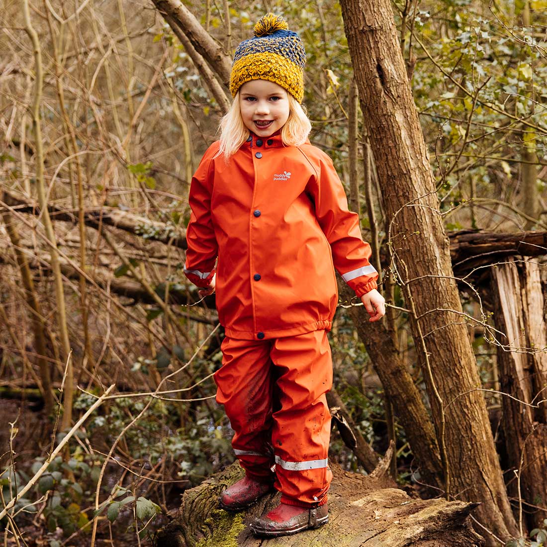 A young child in a Rainy Day Waterproof Jacket and Dungarees Set Red, standing on a log in a woodland setting, wearing a woolly hat.