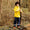 A young boy wearing Muddy Puddles Children's Waterproofs, consisting of a bright yellow raincoat and dark blue waterproof trousers, stands in a woodland.