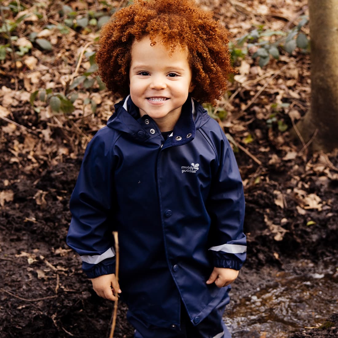 A young child smiling and wearing a Rainy Day Waterproof Jacket Navy in a woodland setting.