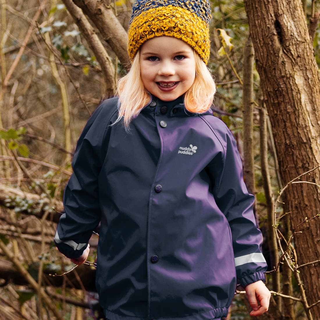A little girl in a wood wearing a navy Rainy Day Waterproof Jacket and knitted hat, smiling at the camera.