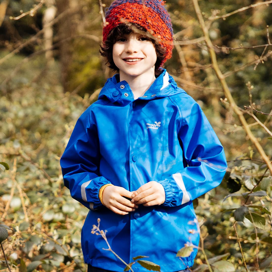 A child wearing a Rainy Day Waterproof Jacket Blue in a wooded area, smiling at the camera.