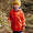 A boy wearing a red Rainy Day Waterproof Jacket standing in a wooded area smiling at the camera. He also has on a yellow and blue bobble hat.