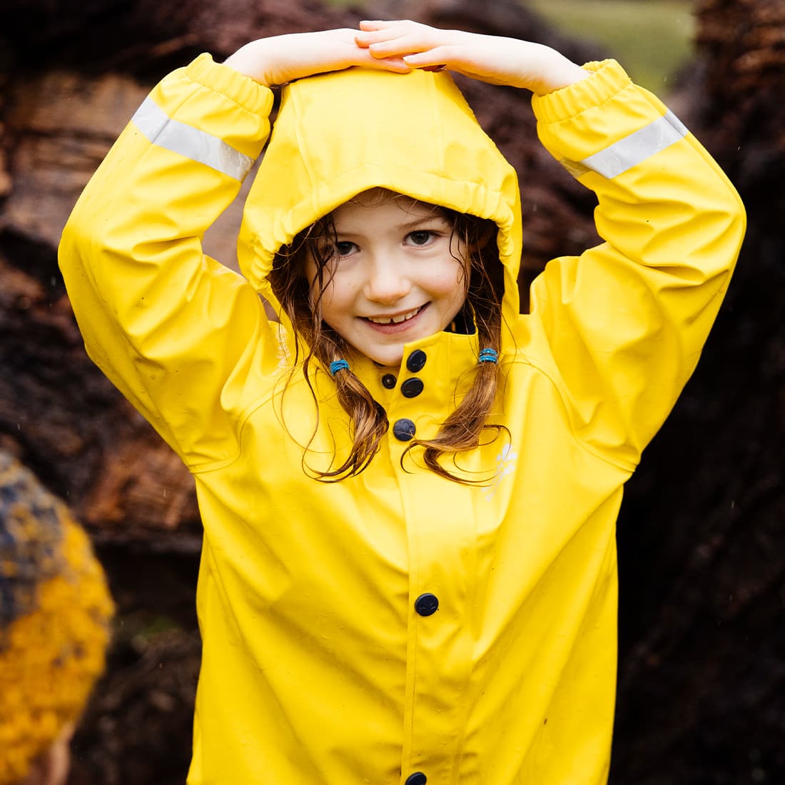 A young girl in a Rainy Day Waterproof Jacket Yellow smiles, holding her hands above her hooded head in a playful manner.