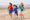 UV Fabrics: Three children run along a sandy beach, holding hands. The kids wear brightly patterned UV protective swimwear and sun hats.