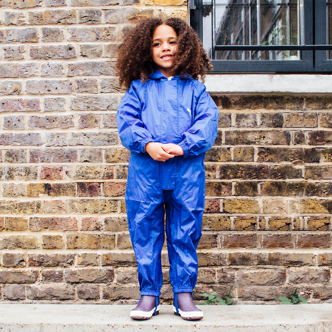 A child wearing a blue Originals Waterproof Puddle Suit, standing in front of a brick wall with a window.
