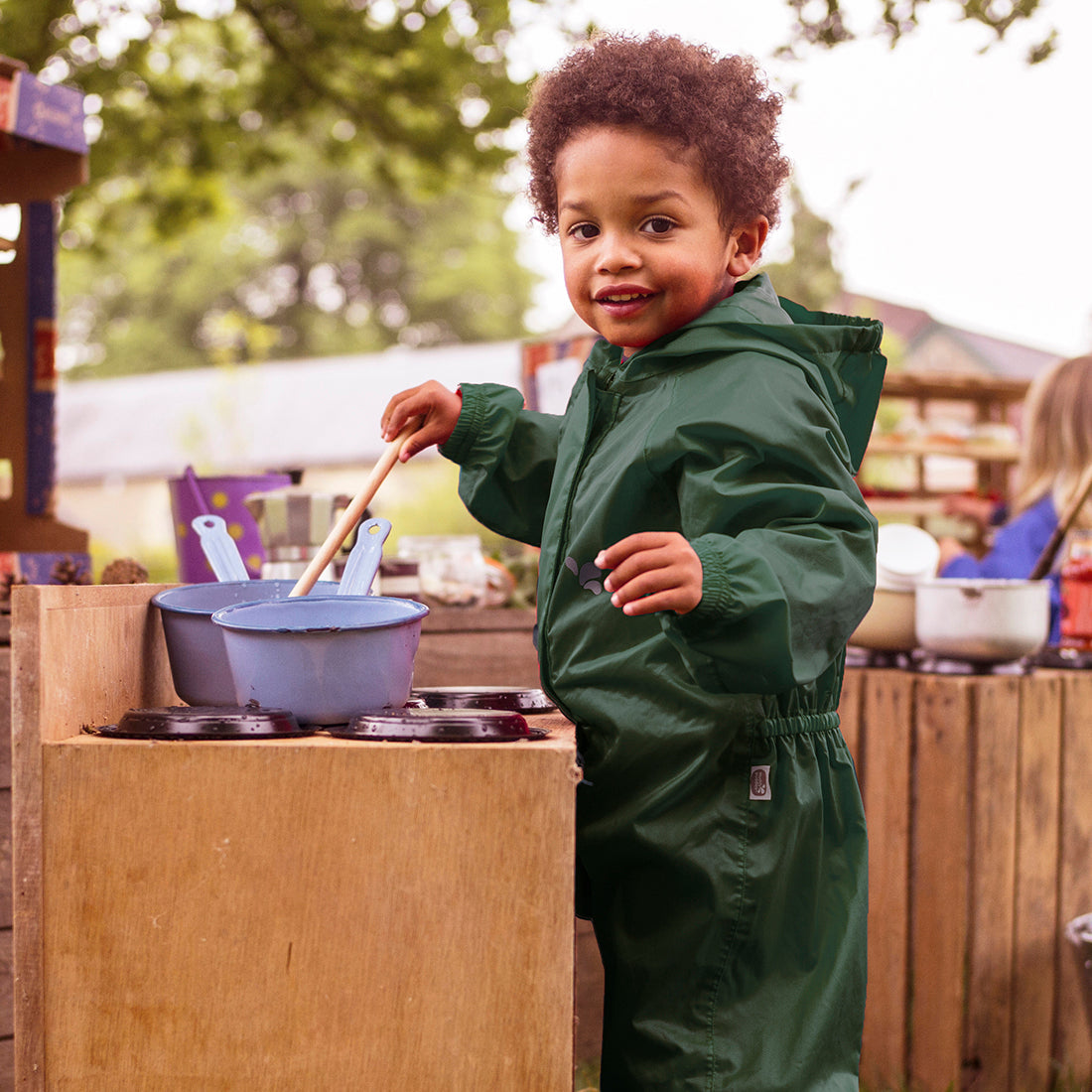 A child in the Originals Waterproof Puddle Suit Green, playing with pots and pans in an outdoor kitchen area.