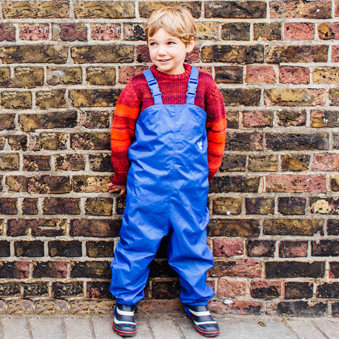 A young child in front of a brick wall wearing Originals Waterproof Dungarees Blue and a red jumper, looking to the right.