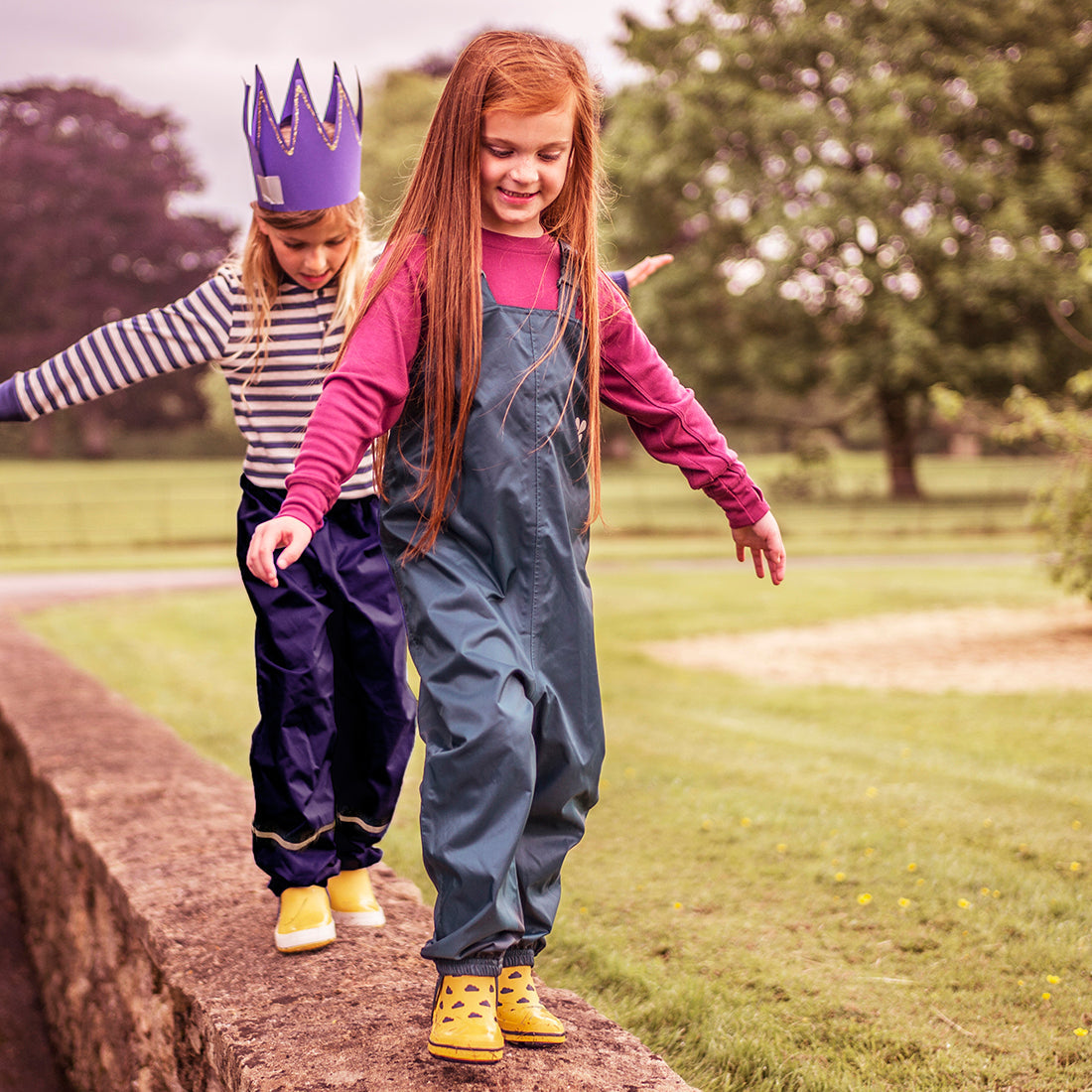 Two young girls balancing on a wall in a park. The girl in front wears green Originals Waterproof Dungarees and yellow boots.