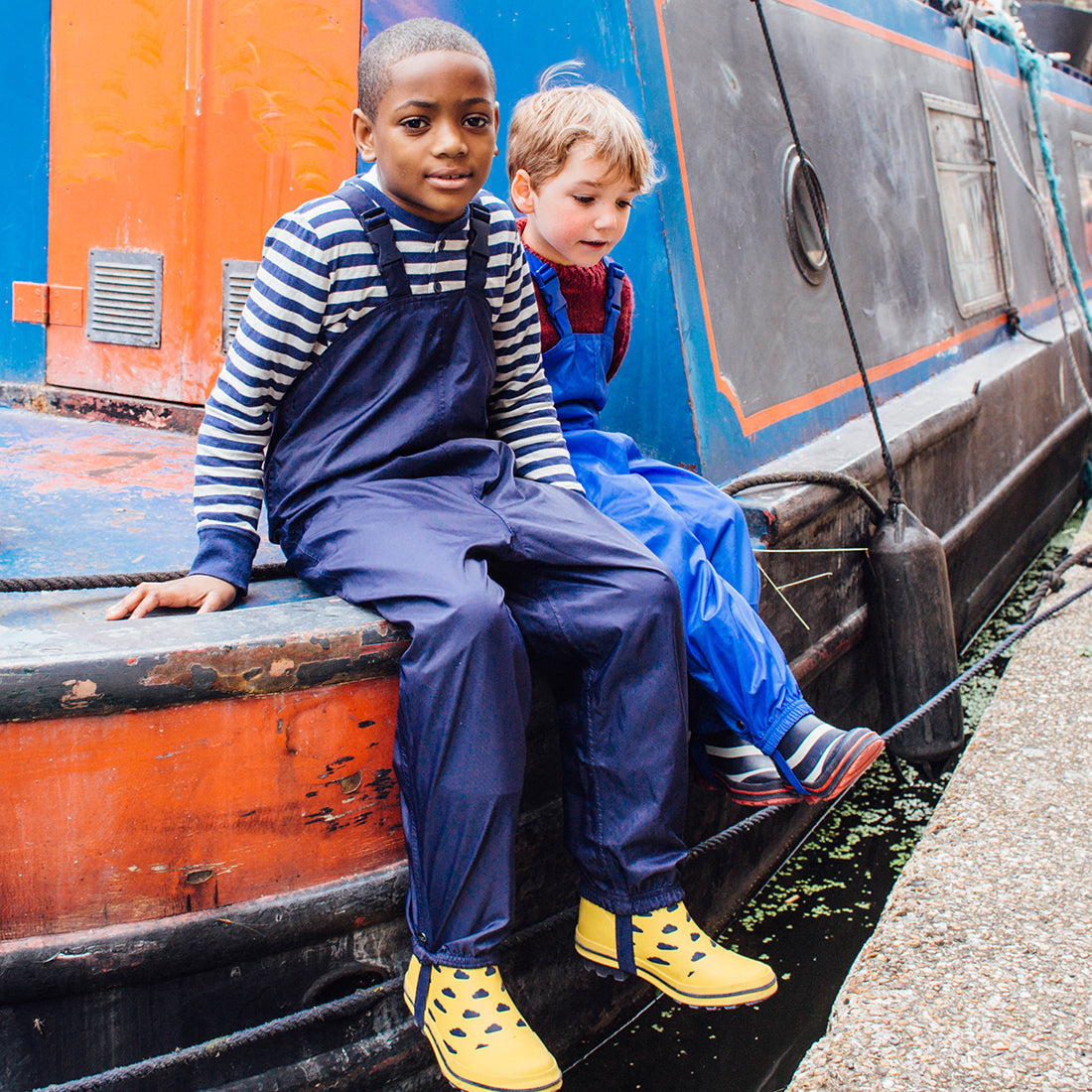 Two boys wearing waterproof dungarees. One boy is wearing navy Original waterproof dungarees, the other is in blue dungarees.