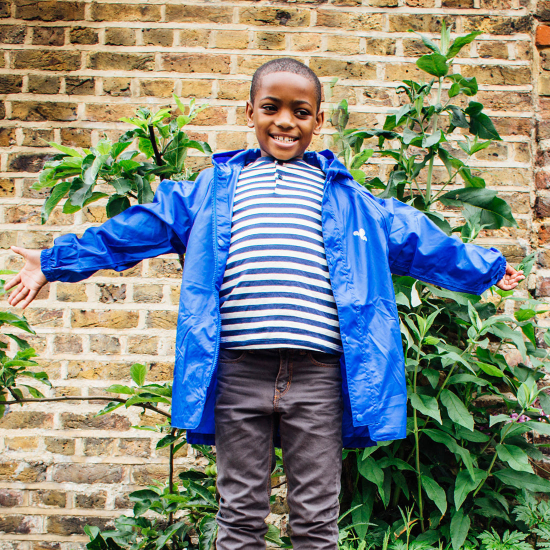 A smiling boy in a garden wears an Originals Waterproof Jacket Blue, with plants and a brick wall in the background.