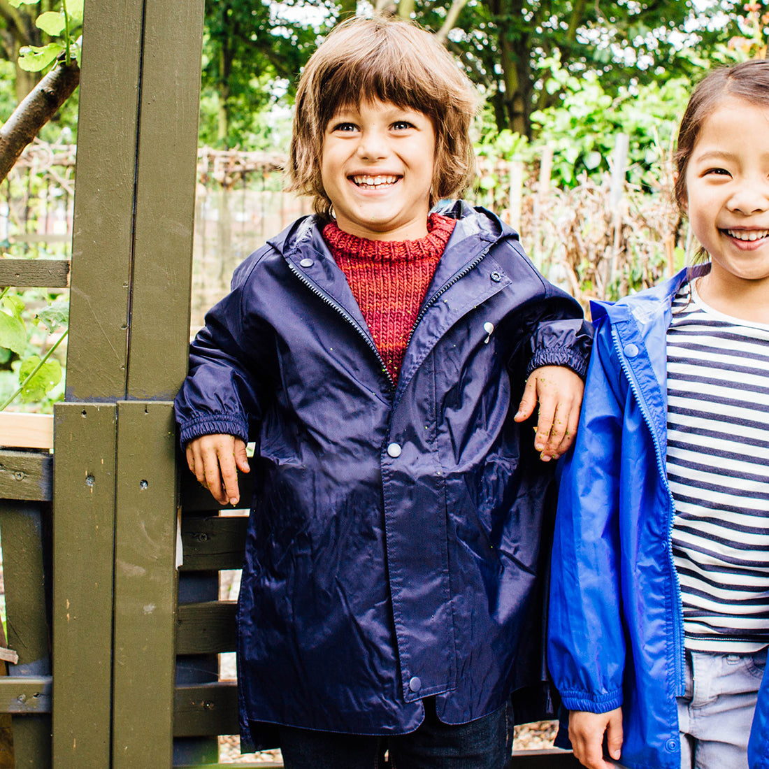 A smiling child in a Originals Waterproof Jacket Navy stands by a wooden fence, with another child partially visible to the right.