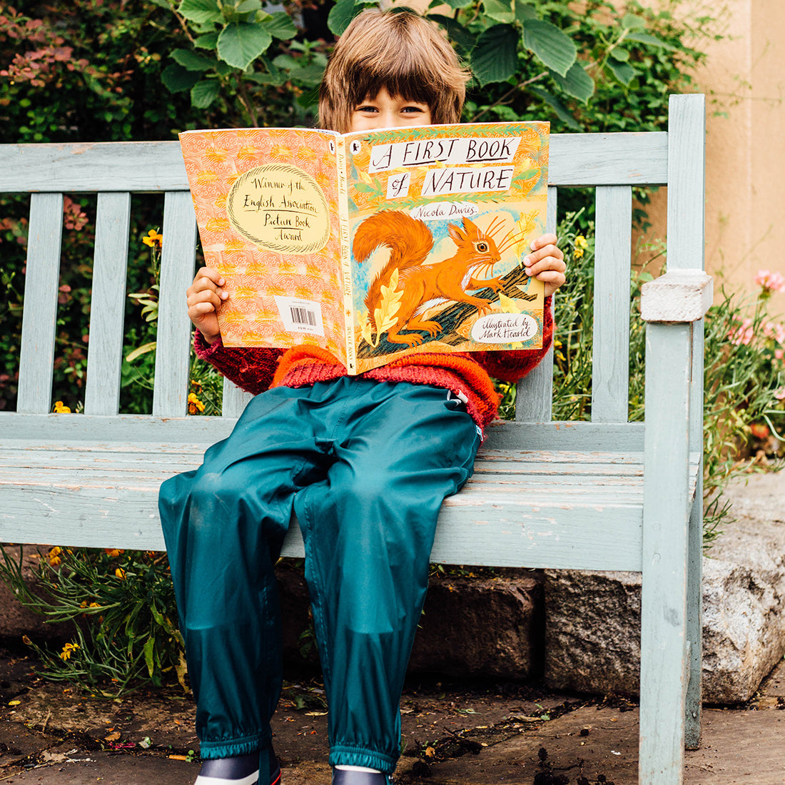 A young child sits on a bench, reading 'A First Book of Nature', wearing Originals Waterproof Trousers Green. Outdoors amongst plants.