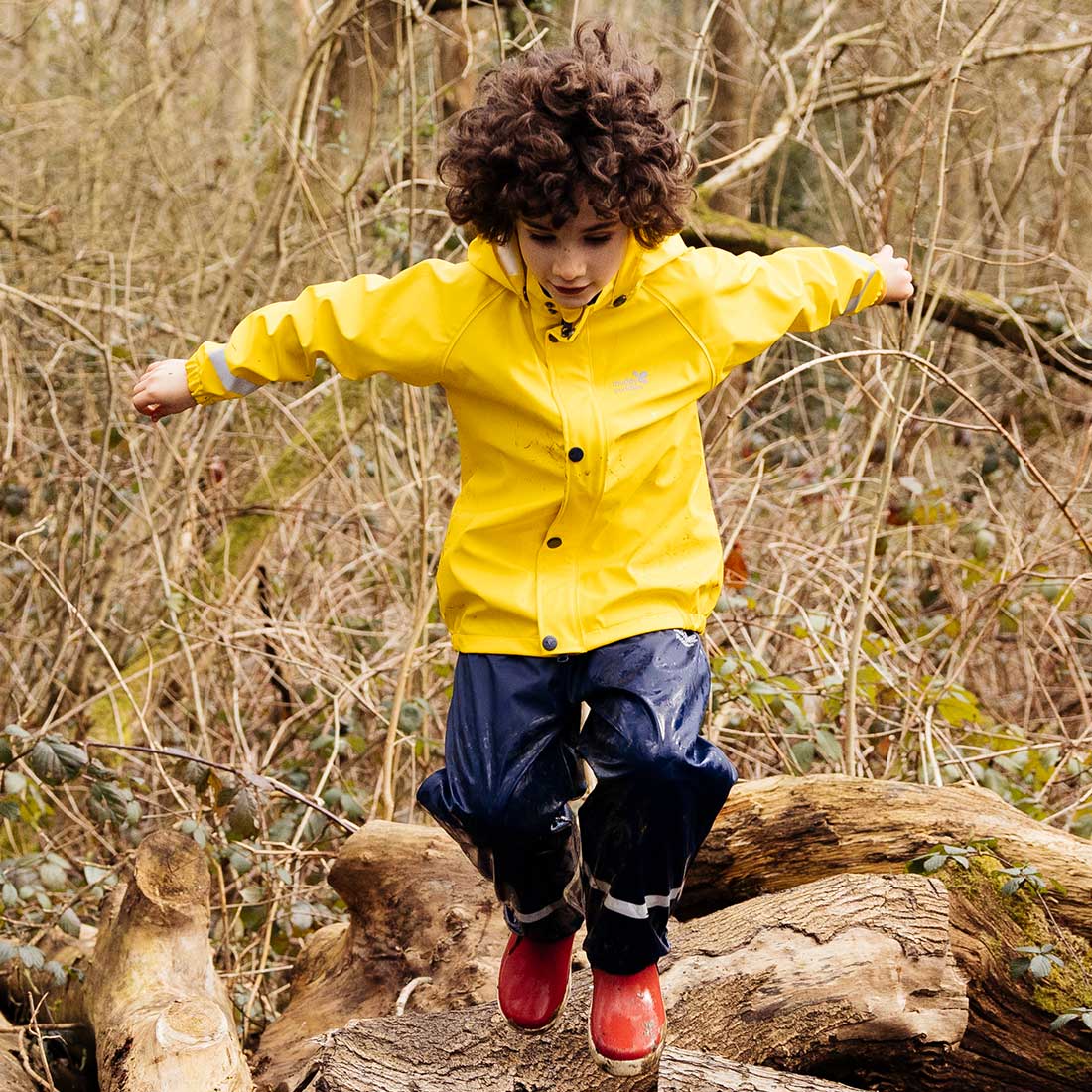 A child wearing a yellow Rainy Day Waterproof Jacket and Dungarees Set, jumping across logs in a forest.