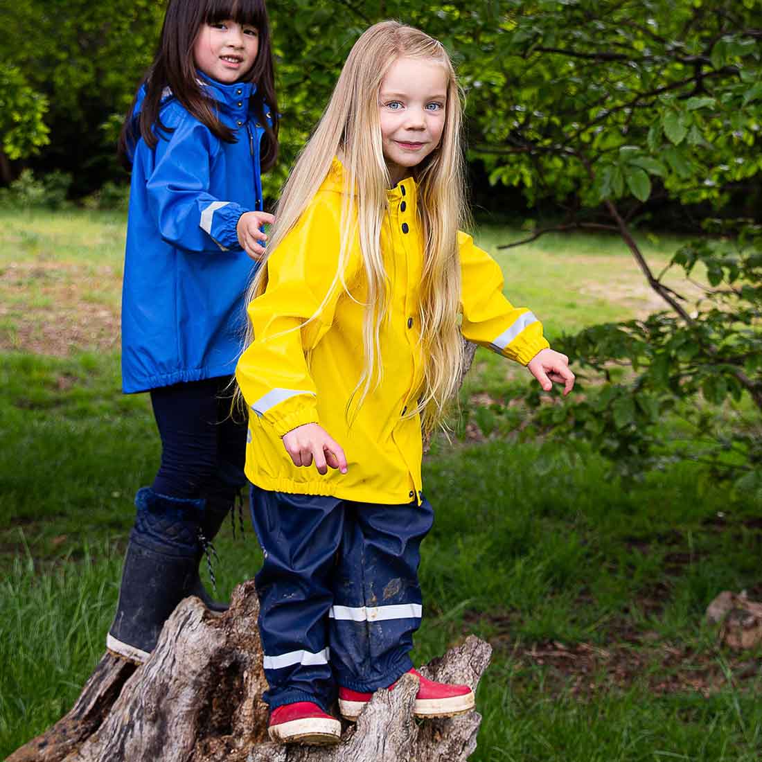 Two girls wearing colourful rainwear, including the 'Rainy Day Waterproof Jacket and Trousers Set Yellow', are on a log in a green woodland area.