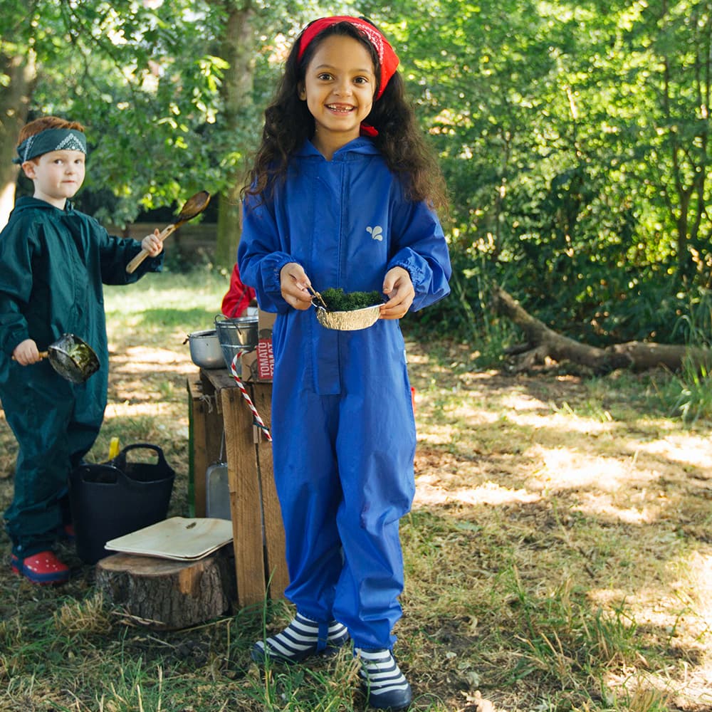 A girl in a blue Originals Waterproof Puddle Suit smiles, holding a dish with greenery, whilst outdoors. Another child stands behind her.