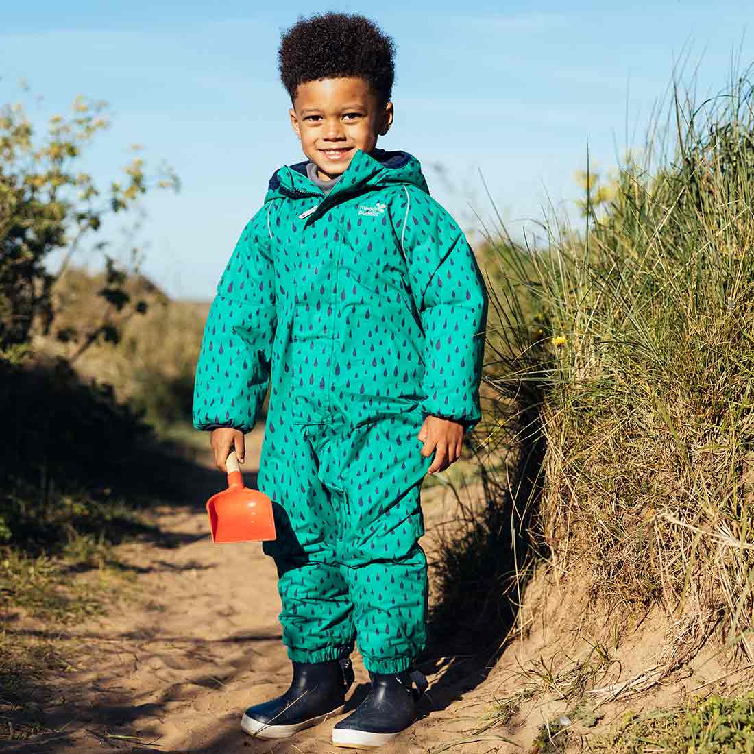 A young boy standing on a sandy path, wearing an EcoWarm Waterproof Snowsuit Green, holding a red shovel near a grassy area.