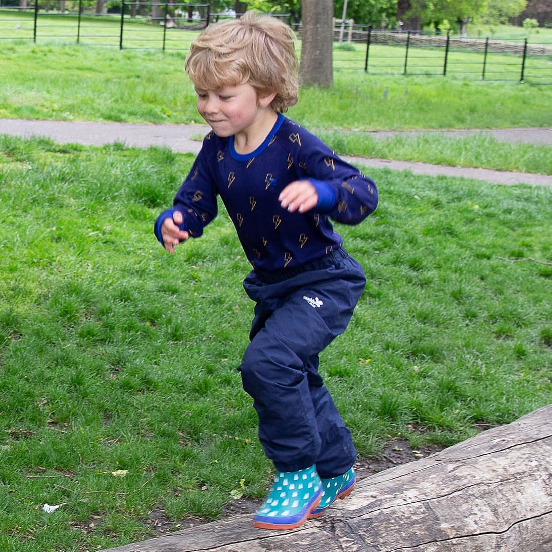 Boy balancing on a log wearing EcoSplash Waterproof Fleece Lined Trousers in navy, paired with teal rain boots, showcasing durable outdoor clothing from Muddy Puddles Children's Waterproofs.
