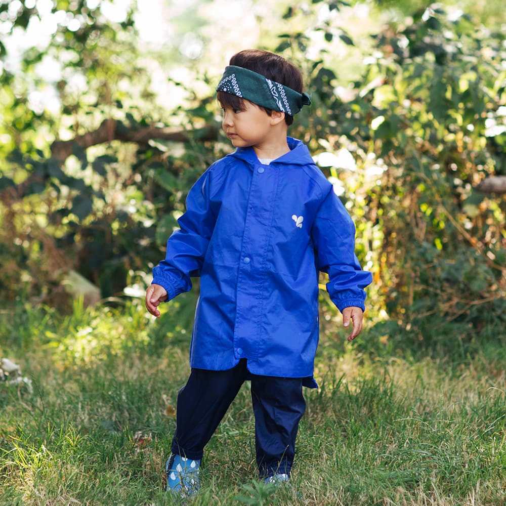 A young child in a garden wearing the Originals Waterproof Jacket Blue, with a bandana on their head and patterned shoes.