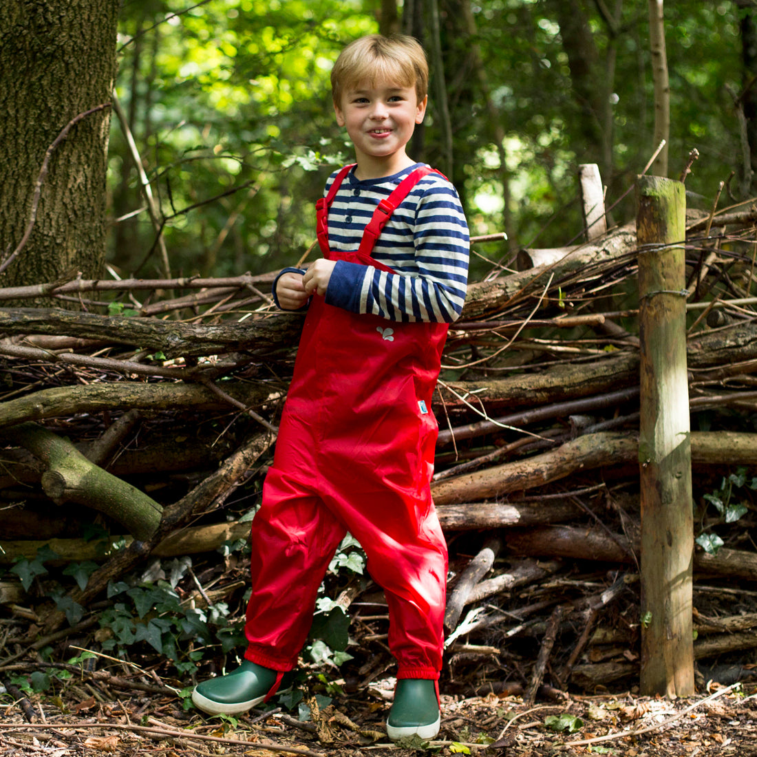 A boy wearing Red Originals Waterproof Dungarees stands outside in a wooded area, in front of a fence made of branches.
