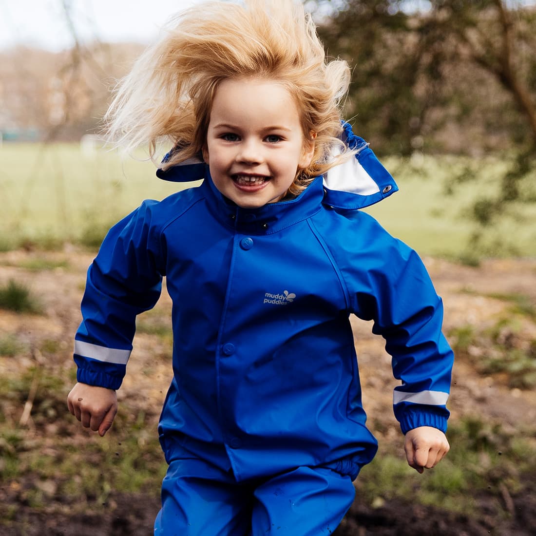 A smiling child wears a blue Rainy Day Waterproof Jacket while running outdoors, blonde hair flowing and creating a joyful scene.
