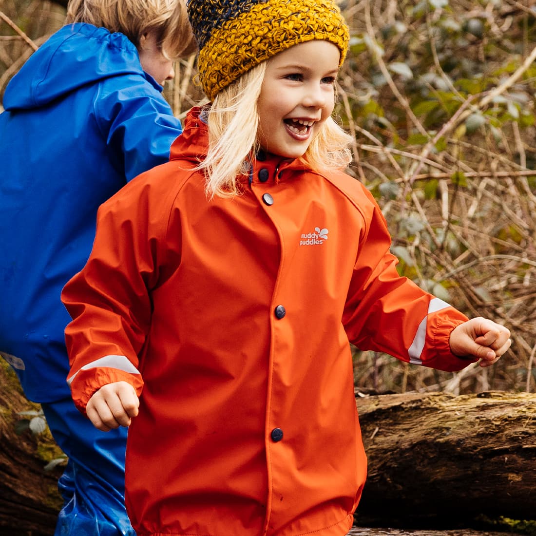 A happy child in an orange waterproof jacket with Muddy Puddles logo, wearing a striped hat, outdoors with another child in a blue jacket.