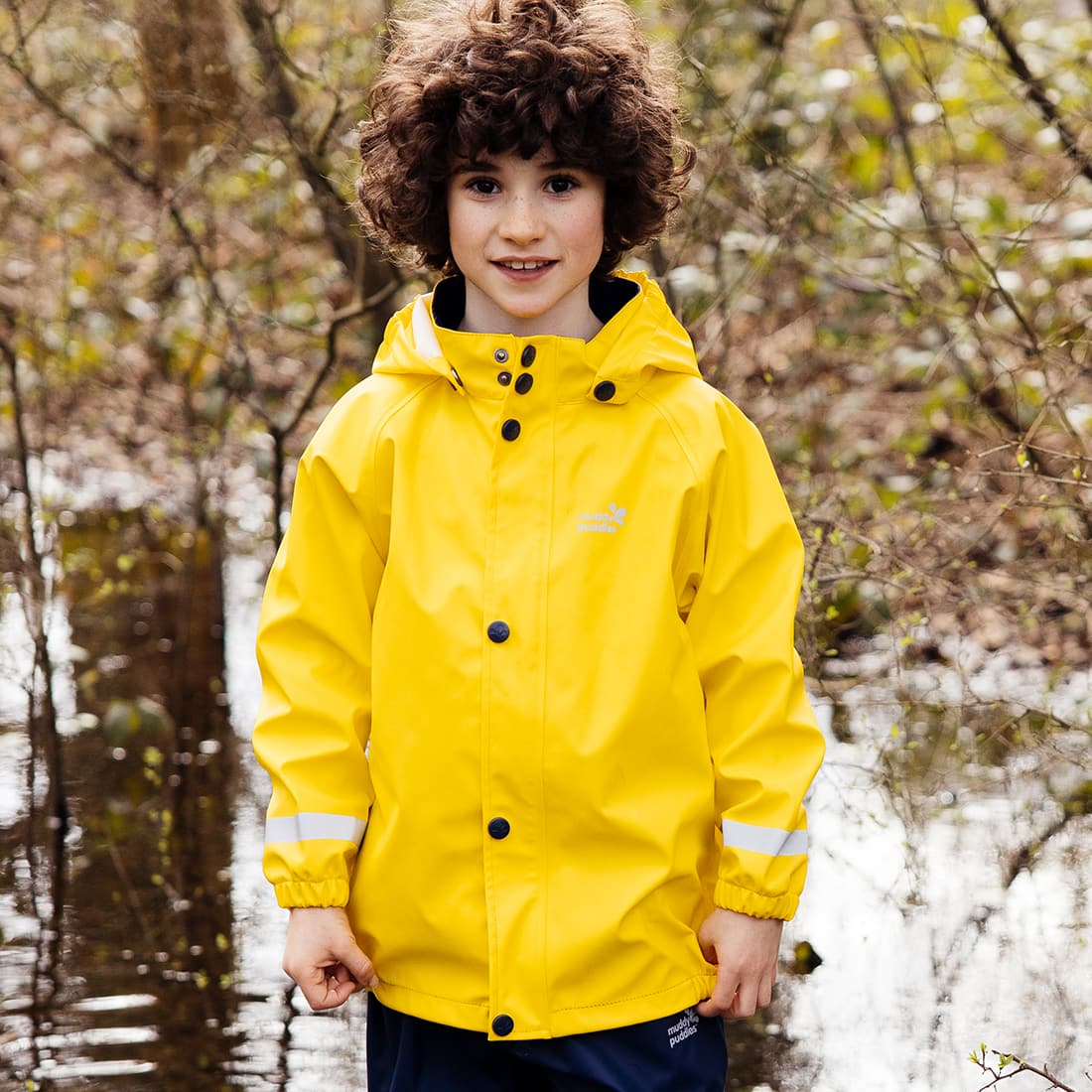 A boy in a forest wearing a Rainy Day Waterproof Jacket Yellow. He has curly hair and is smiling for the camera.