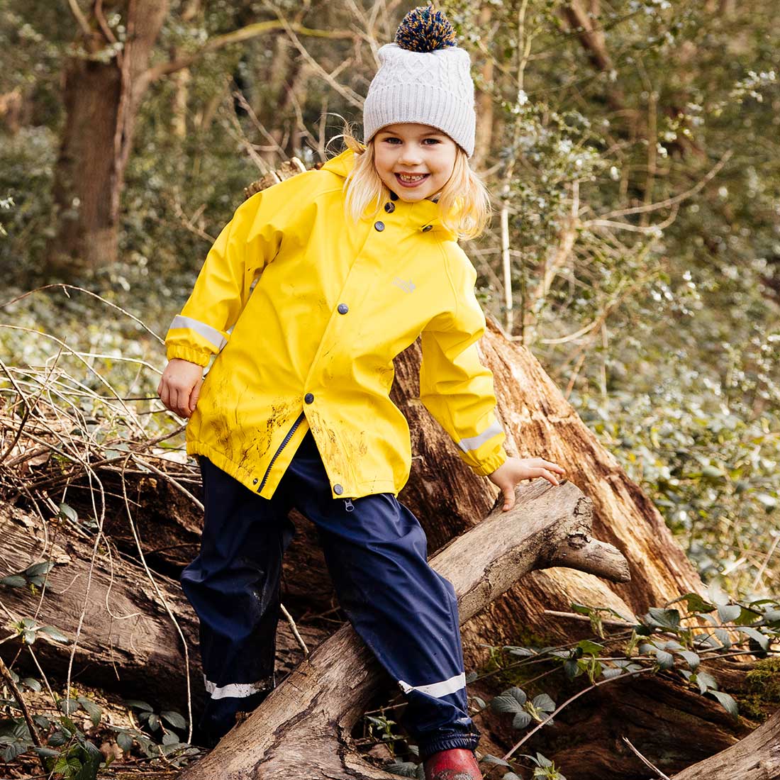 A girl in a Rainy Day Waterproof Jacket and Dungarees Set Yellow, climbing on a log in a forest, smiling at the camera.