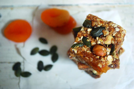 granola bars on a white surface, next to dried apricots and sunflower seeds