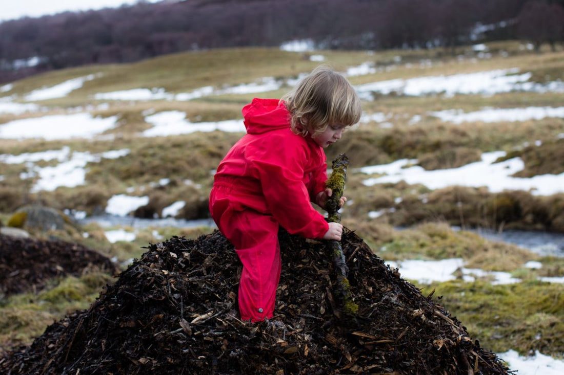 boy standing on pile of bark holding a large stick, wearing red muddy puddles puddlesuit