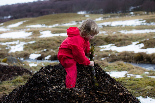 boy standing on pile of bark holding a large stick, wearing red muddy puddles puddlesuit