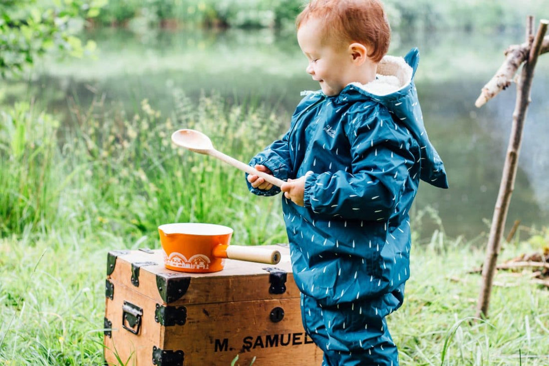 toddler playing by a pond, wearing muddy puddles scampsuit, holding a wooden spoon, with an orange pan and a wooden trunk in the background