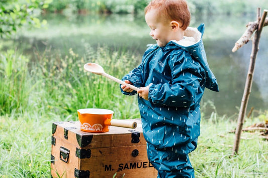 toddler playing by a pond, wearing muddy puddles scampsuit, holding a wooden spoon, with an orange pan and a wooden trunk in the background