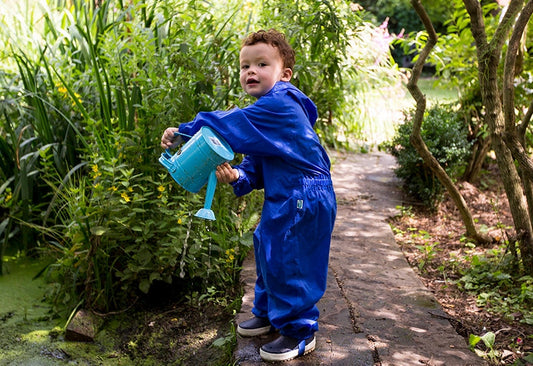 toddler boy emptying a blue watering can into a small pond, wearing a bold blue muddy puddles puddlesuit