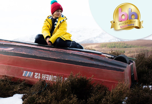 child sitting on overturned boat wearing yellow waterproof jacket and red bobble hat, with bizziebaby logo in upper right corner