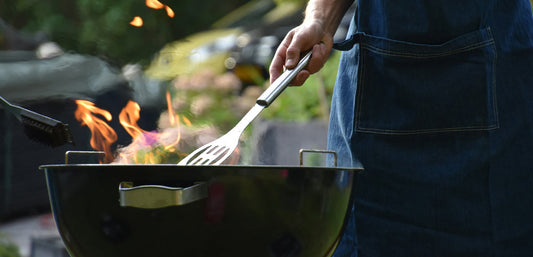 Person grilling on outdoor barbecue with flames, wearing blue apron, summer cookout scene