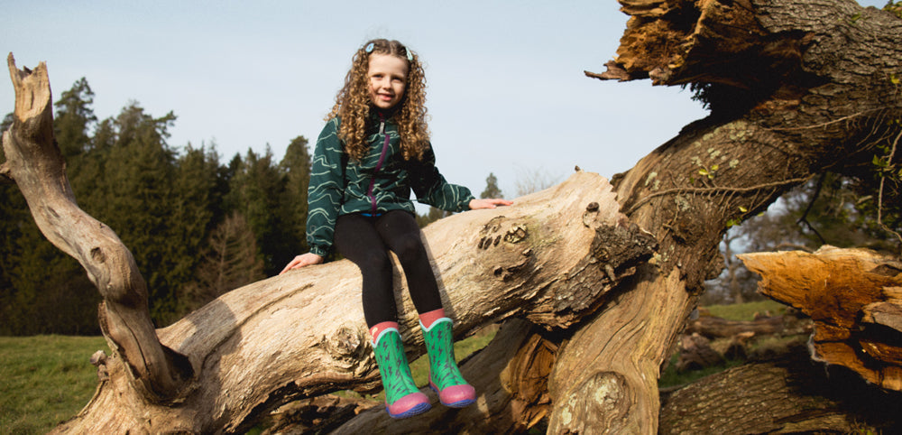 girl sitting on fallen tree, wearing muddy puddles waterproof jacket and boots